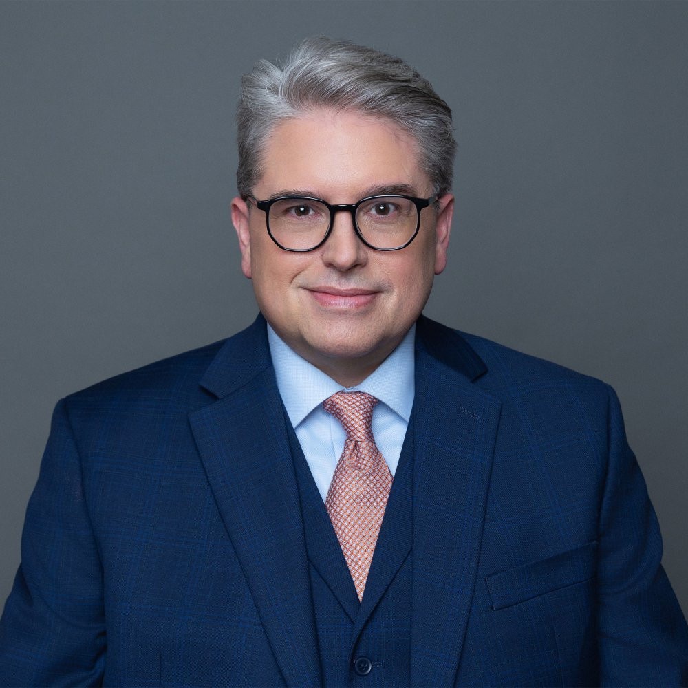 Man with gray hair and glasses wearing a blue suit, light blue shirt, and peach tie, posing against a plain gray background.