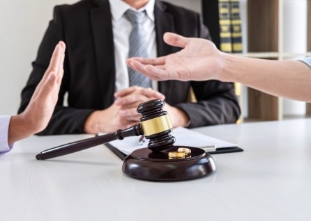 Two people gesture with their hands in disagreement near a gavel and wedding rings, while a houston family law attorney in a suit sits behind them at a desk.