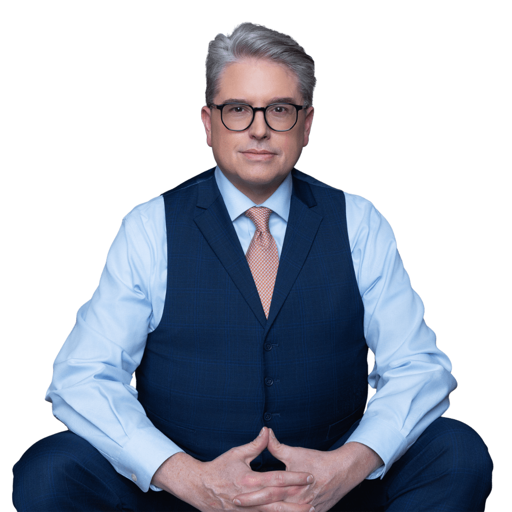 A man with gray hair and glasses sits cross-legged, wearing a blue suit vest, light blue shirt, and a peach tie, against a plain white background—his poised demeanor reminiscent of an expert ready to offer guidance on topics such as those found in a Houston family law blog.